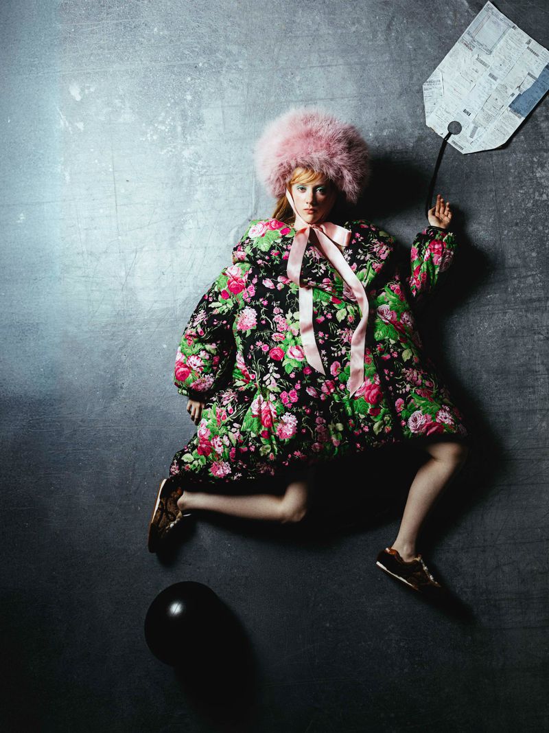 A fashionable woman with a pink fur hat and floral jacket is styled creatively with a paper tag and black balloon on a grey backdrop.