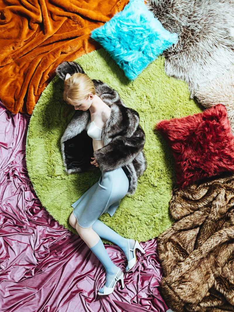 Overhead fashion shot of a woman in a fur coat, light blue dress, and heels lying on a green rug with assorted textured pillows and blankets.