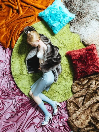 Overhead fashion shot of a woman in a fur coat, light blue dress, and heels lying on a green rug with assorted textured pillows and blankets.