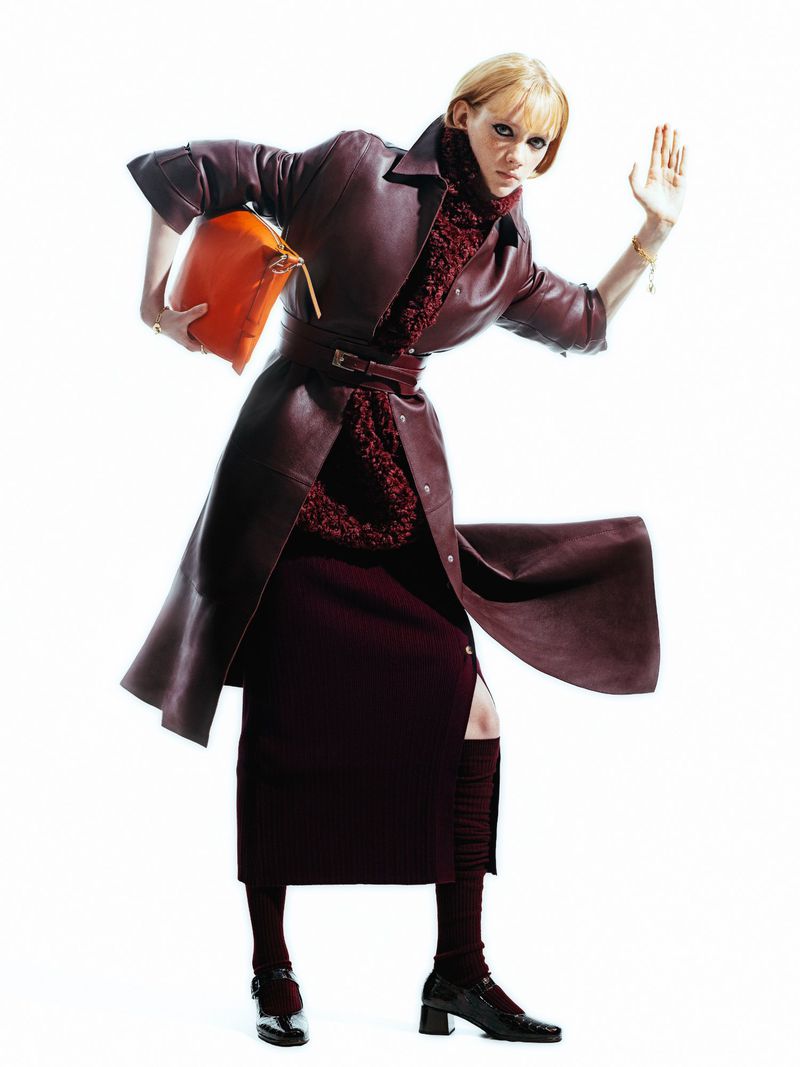 Fashion model in a long leather coat, midi skirt, and block heels, holding an orange purse against a stark white backdrop. Studio shot.