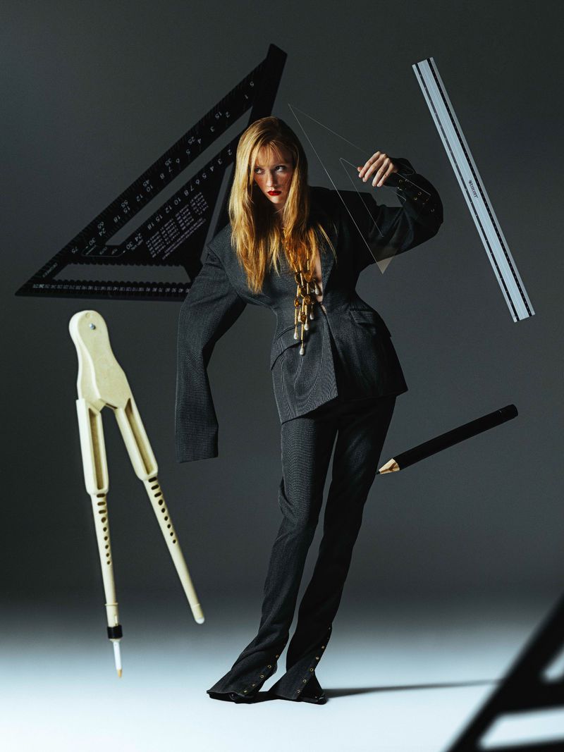 Woman in suit surrounded by drafting tools in studio lighting. She's posing with a clear triangle in her hand.