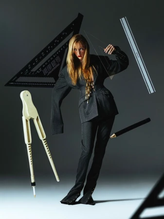 Woman in suit surrounded by drafting tools in studio lighting. She's posing with a clear triangle in her hand.