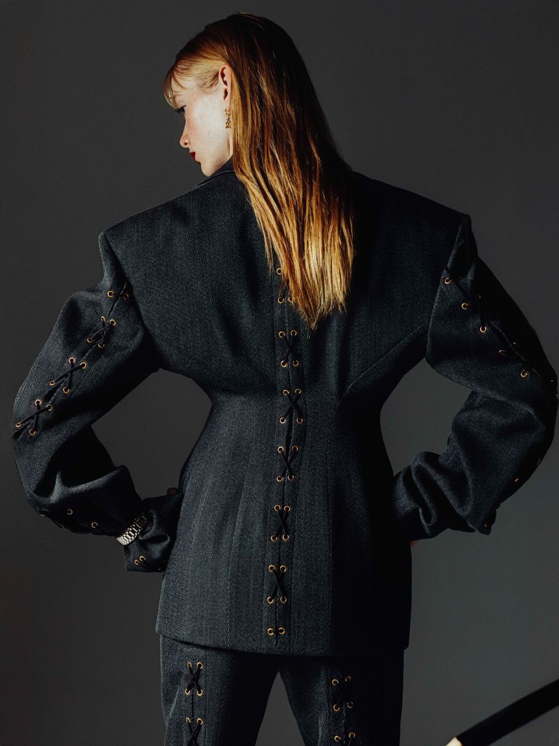 A woman wearing a denim jacket and pants, with lace-up details on sleeves and back, poses against a gray backdrop, showcasing fashion design.