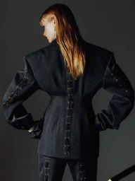 Model seen from behind wearing a dark grey structured jacket with lace-up details against a dark background