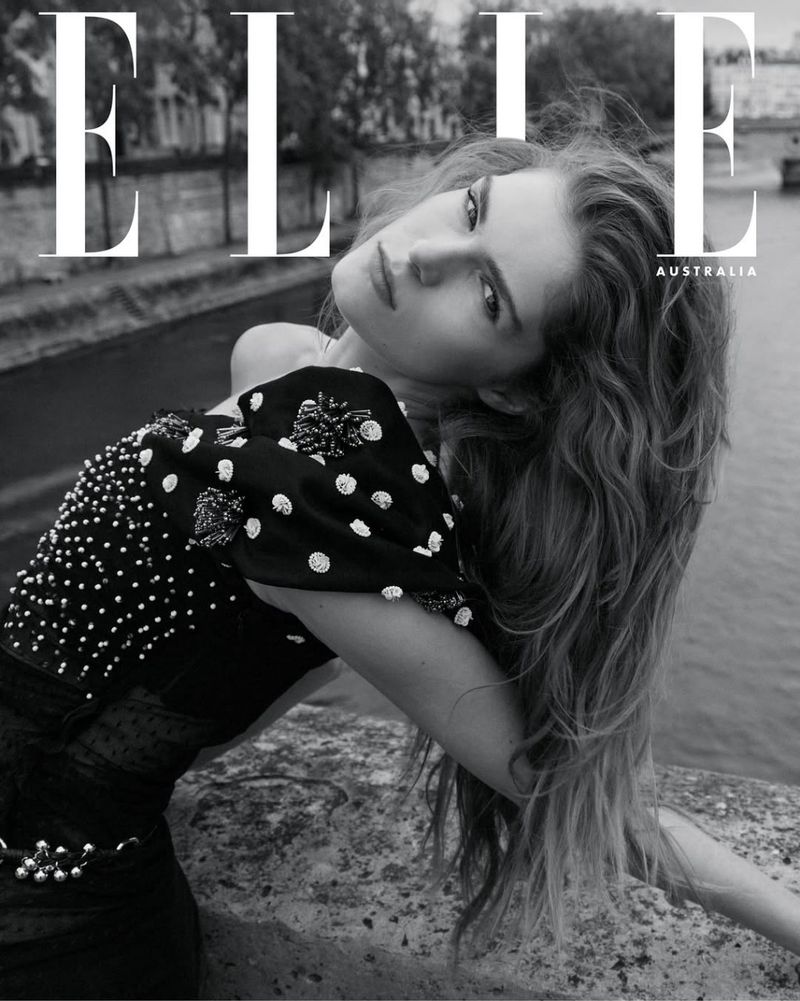 Black and white magazine cover of a woman with long hair, wearing a polka dot top, posing on a stone bridge over a river.