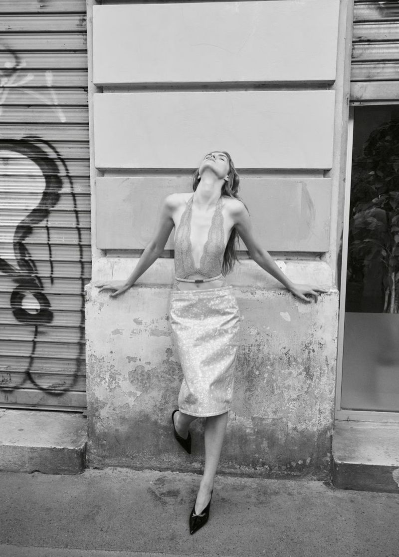 Black-and-white outdoor fashion shot of a woman in a halter top and skirt leaning against a building with graffiti, eyes closed, enjoying sunlight.