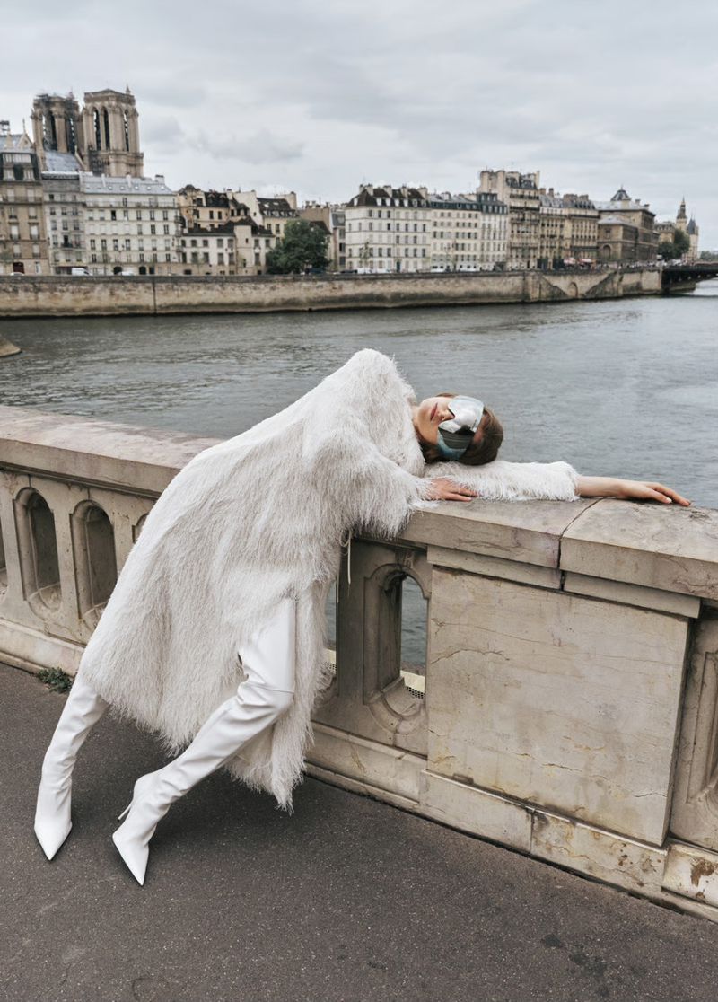Fashion shoot of a woman in a white coat and boots reclining on a stone bridge with a city and a river in the background.