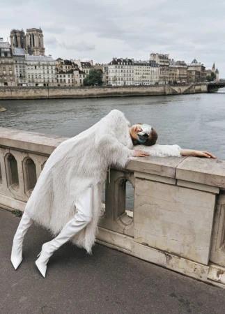 Fashion shoot of a woman in a white coat and boots reclining on a stone bridge with a city and a river in the background.