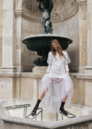 Fashionable woman in a white dress and black sandals stands on metal bars in a stone fountain with a statue in the background.