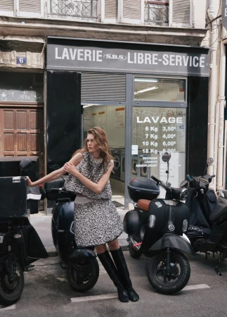 A fashionable woman poses next to black scooters in front of a laundromat in an urban setting, wearing a textured top and skirt with knee-high boots.