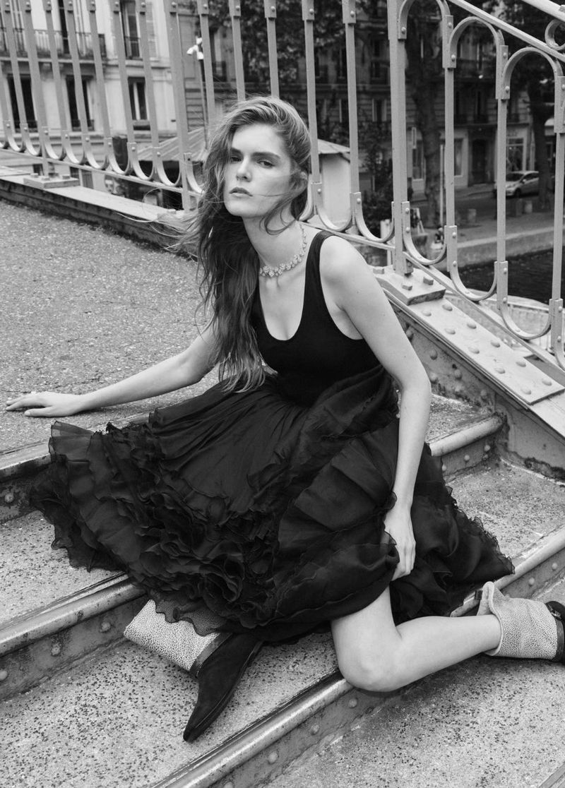 Black and white fashion shot of a woman in a black dress and boots kneeling on metal steps, outdoor urban setting with trees.