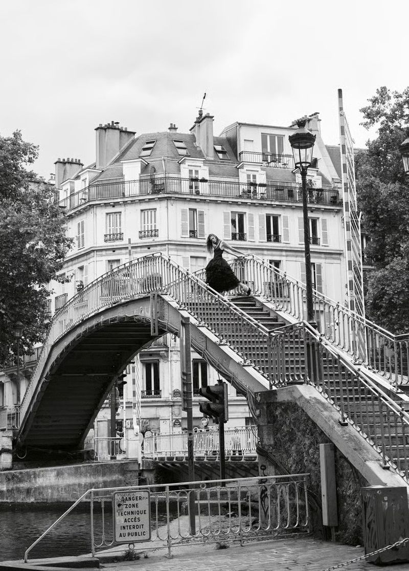 Model Anna Robinson poses in a black dress atop an arched footbridge over a canal with classic Parisian buildings behind her.
