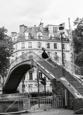 Model Anna Robinson poses in a black dress atop an arched footbridge over a canal with classic Parisian buildings behind her.