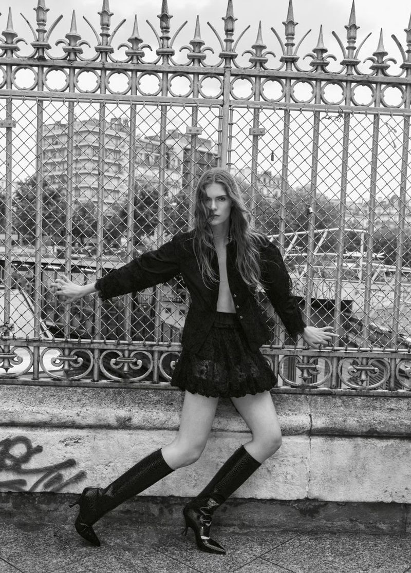 Fashion editorial shot in black and white featuring a long-haired woman in a short skirt and boots, posing in front of an ornate fence.