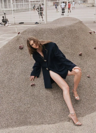 Woman in a dark blue jacket and high heels poses on a textured, tan-colored slope with a fenced court and other people visible in the background.
