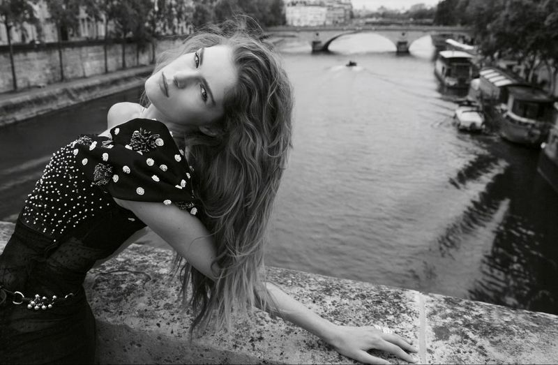 Black and white portrait of a fashion model leaning on a bridge over a river with buildings and bridge in the background.