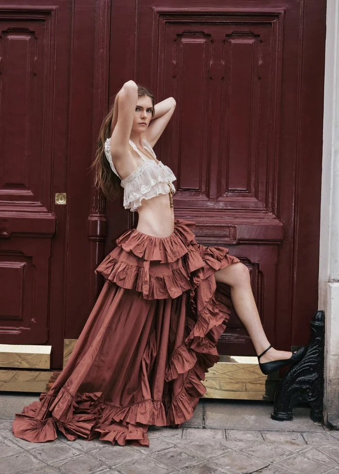 Model wearing a ruffled terracotta maxi skirt and white crop top posing against a large dark red wooden double door