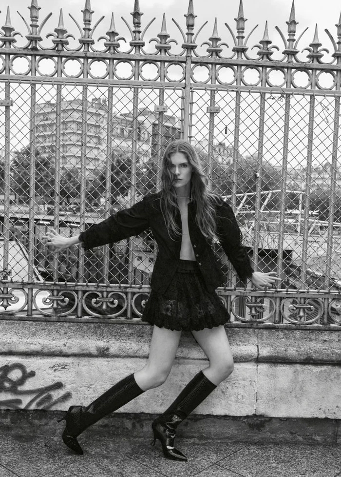 Model Anna Robinson poses in a black jacket, ruffled miniskirt, and heeled boots against an ornate wrought iron fence in black and white.