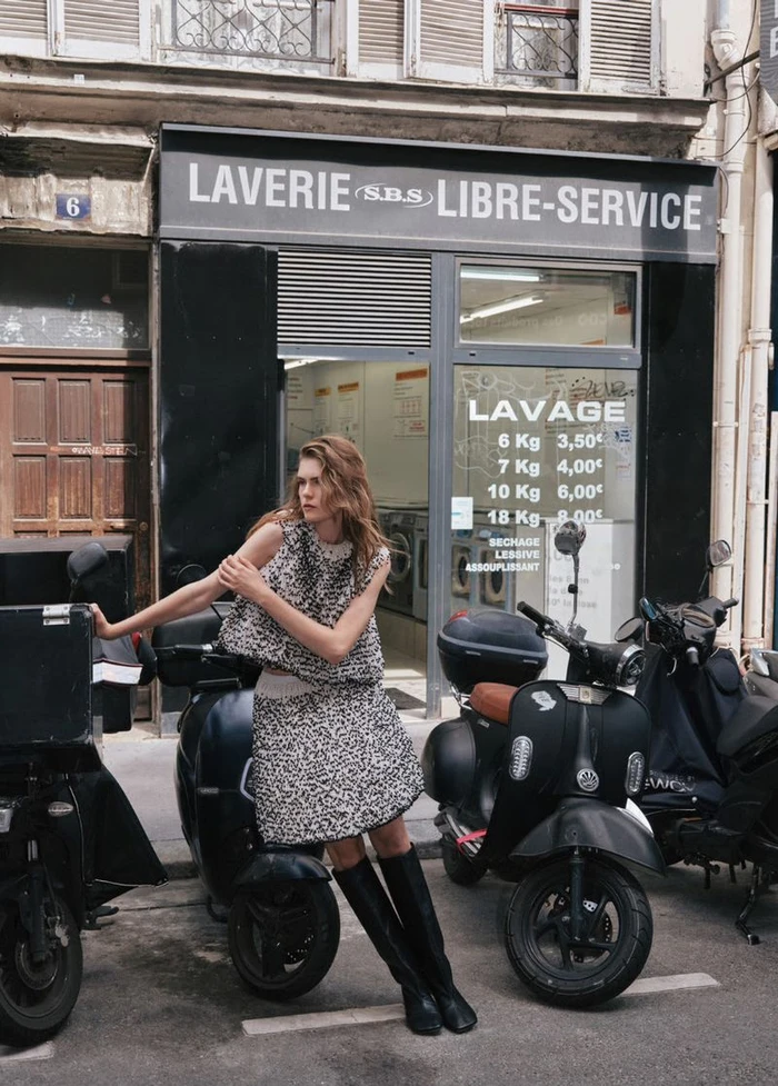 Model standing on a street next to scooters in front of a laundromat wearing a patterned two-piece skirt set and tall black boots.