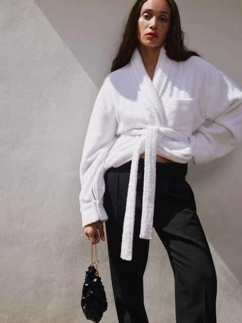 A fashionable woman in a white robe and black pants poses against a light gray wall, accessorized with a small black purse.