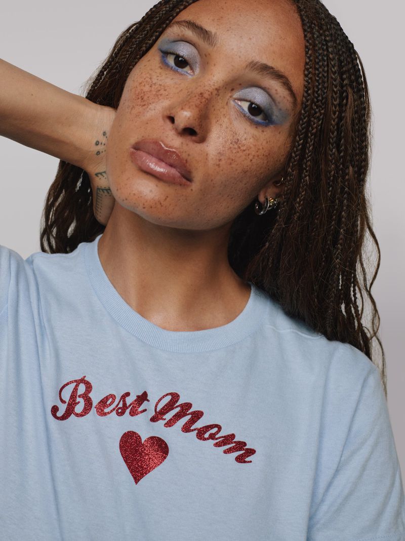 Woman with freckles, wearing a light blue T-shirt with a glittery 'Best Mom' graphic design, poses in front of a neutral background.