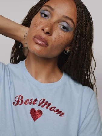 Woman with freckles, wearing a light blue T-shirt with a glittery 'Best Mom' graphic design, poses in front of a neutral background.