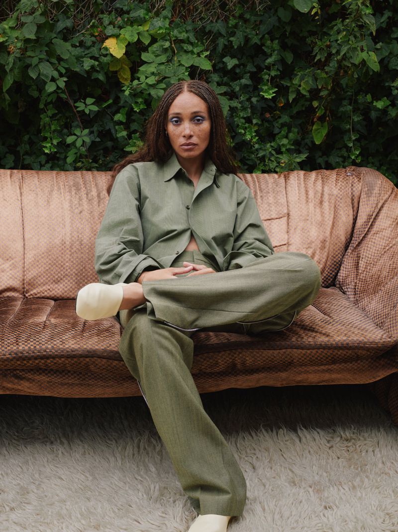 An adult female with braids poses in green clothing on a brown couch with green foliage behind, illuminated by soft natural light.