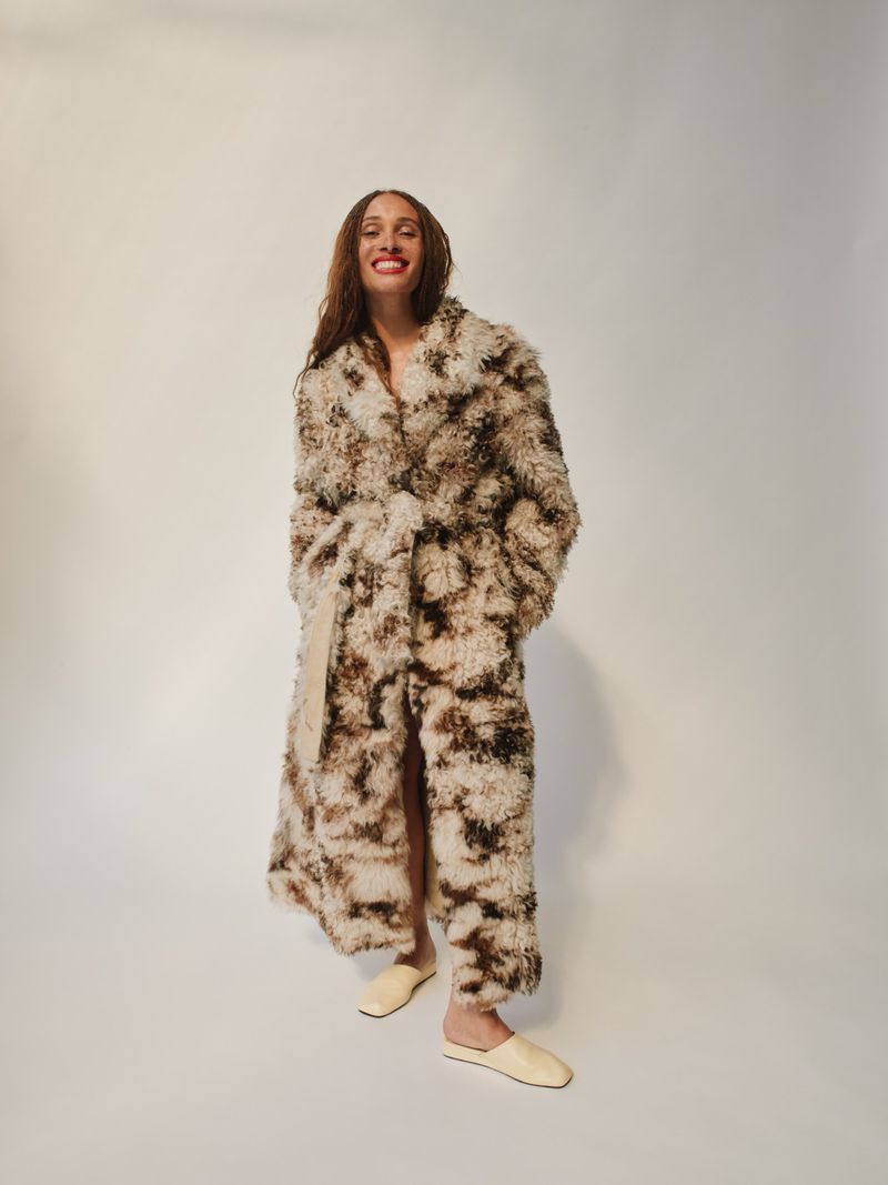 A woman in a cream and brown patterned faux fur coat stands in front of a white backdrop, smiling and looking at the camera.