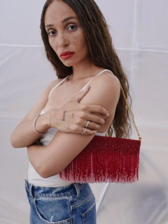 Adult female with long braided hair, wearing a white tank top and blue jeans, posing with a red beaded clutch in a photo studio.