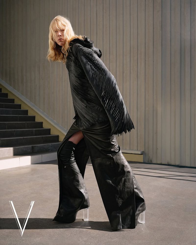 Fashion editorial shot of a blonde female model in dramatic black clothing posing against a concrete wall with stairs in the background.