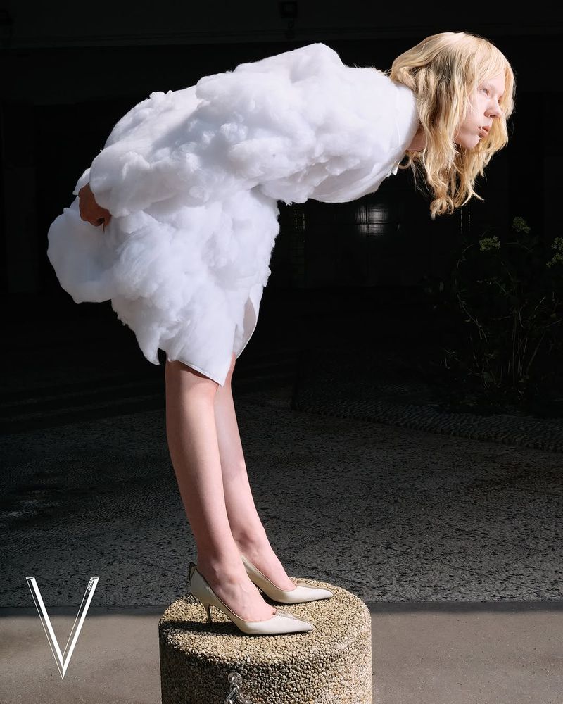 Fashion editorial shot of a fair-skinned blonde woman in a puffy white dress standing on a small pedestal in high heels.