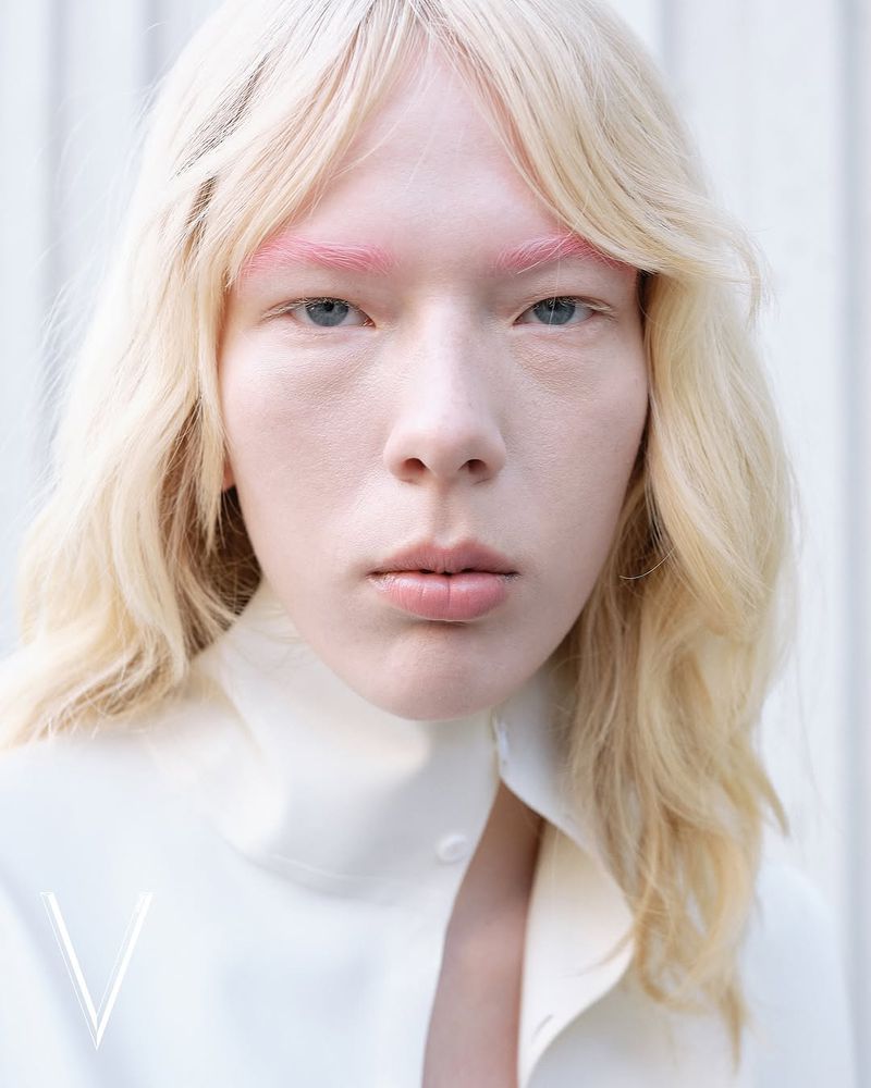Close-up portrait of a person with pink eyebrows and blonde wavy hair, wearing a white collared shirt against a white background.