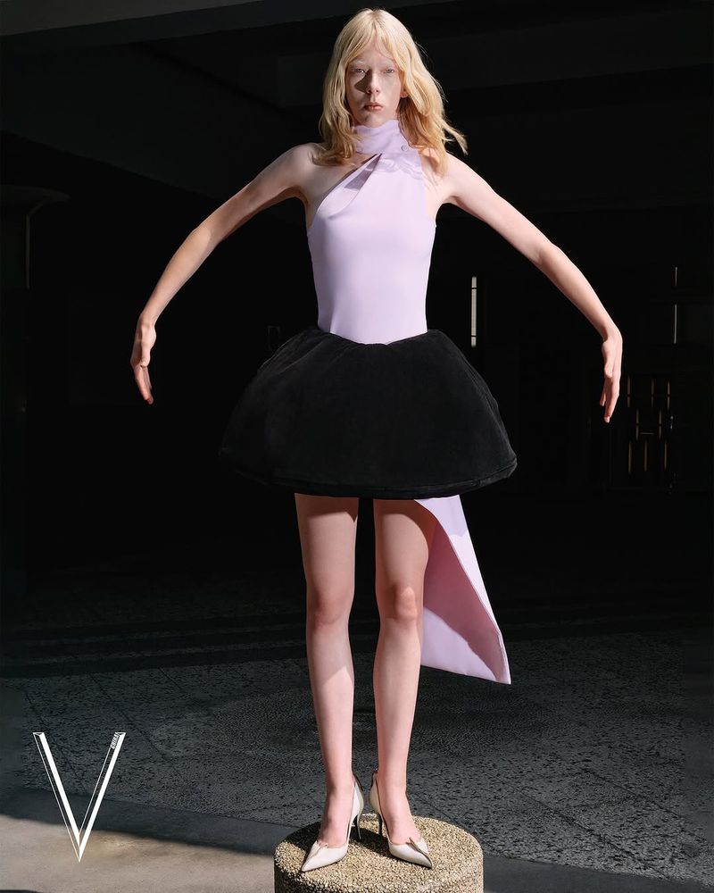A slender model poses on a pedestal in a fashion shot, wearing a lilac bodysuit, black bubble skirt, and white heels against a dark background.