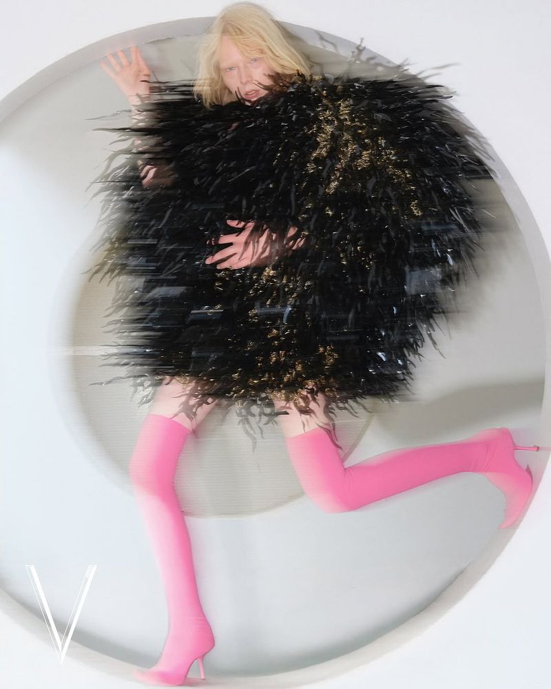 Fashion shot of a blonde female adult in a black feathered jacket and pink boots posing in a blurred white circle, highlighting fashion elements.