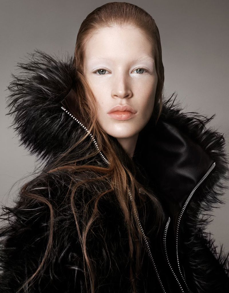 Portrait of a fair-skinned woman with light brown hair wearing a black fur coat, looking directly at the camera against a grey backdrop.