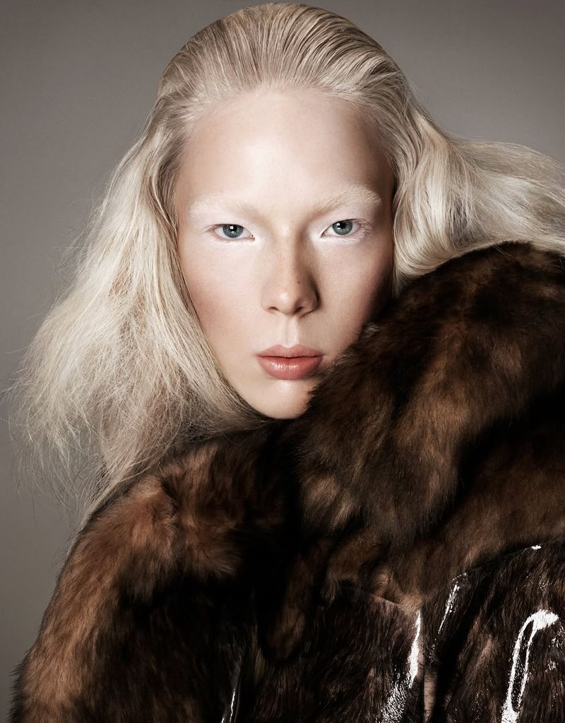A pale-skinned woman with white hair and brows gazes forward, enveloped in a brown fur coat against a neutral gray backdrop in a studio shot.