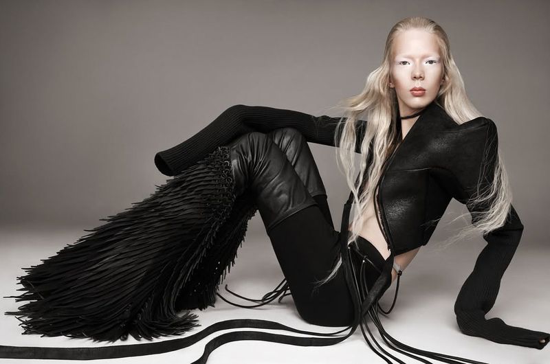 Fashionable albino model poses seated in black clothing, featuring a leather jacket, fringe thigh-high boots against a grey background.