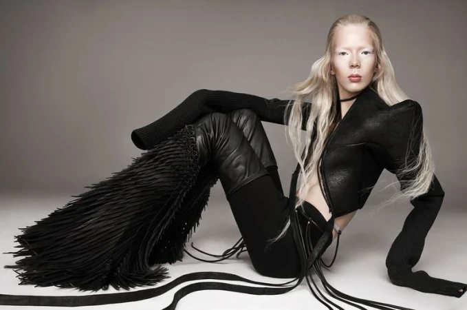 Fashionable albino model poses seated in black clothing, featuring a leather jacket, fringe thigh-high boots against a grey background.