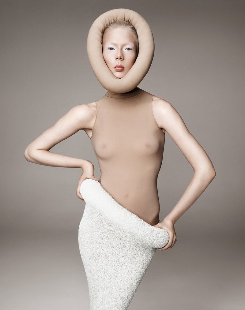 A person poses in a studio with a modern art donut headpiece and matching tan bodysuit and textured material wrapped around the waist.