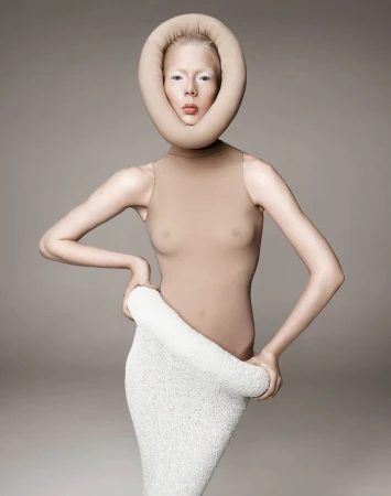 A person poses in a studio with a modern art donut headpiece and matching tan bodysuit and textured material wrapped around the waist.