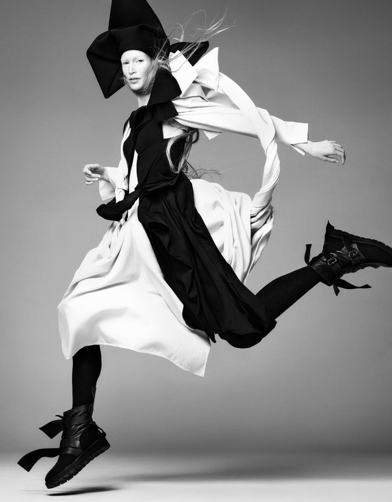 Fashion editorial shot of a person in mid-air wearing a sculptural black-and-white outfit with platform boots against a grey background.