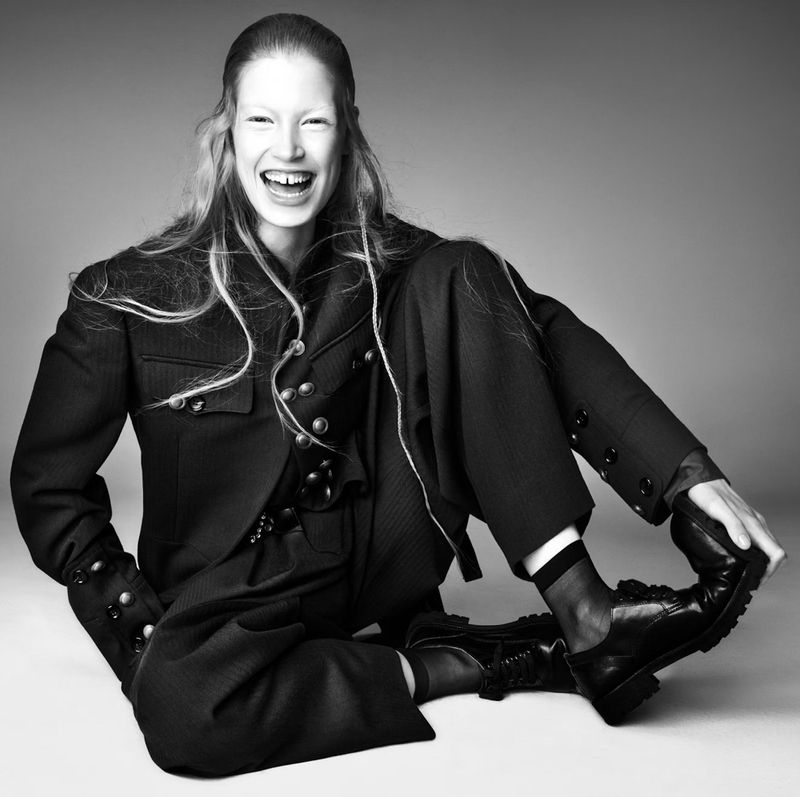 Smiling woman in a black military-style jacket and pants poses in a studio. She is seated on the floor with one leg bent.