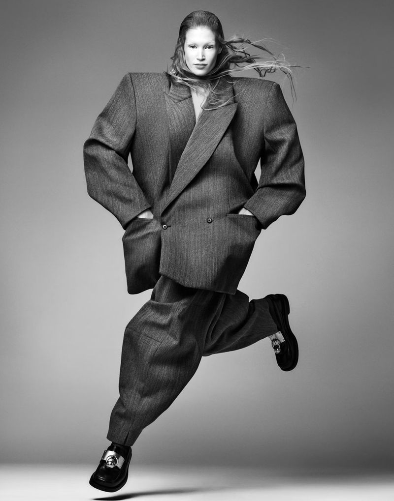 A person wearing an oversized grey suit and black penny loafers, is in a mid-air leaping pose against a neutral grey background.