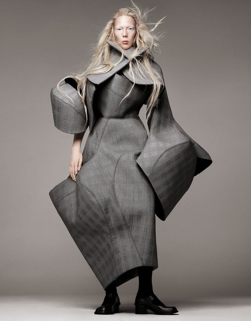 A woman in a gray textured coat poses in a studio. The full shot is taken at eye level. The coat is oversized and has geometric accents.