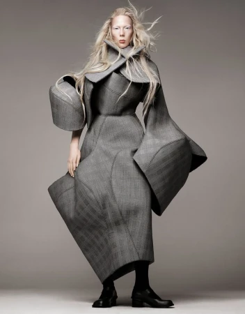 A woman in a gray textured coat poses in a studio. The full shot is taken at eye level. The coat is oversized and has geometric accents.