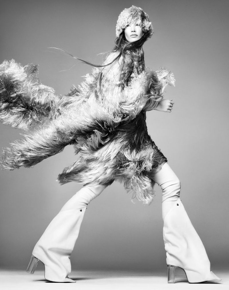 Monochrome fashion shot: a long-haired model wears a feathered dress, feathered hat, and platform boots against a plain background.