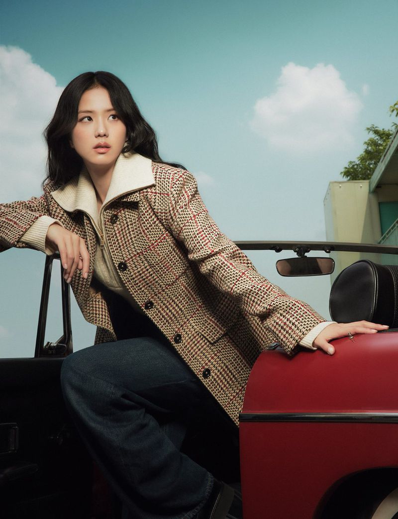A woman with dark hair poses leaning against a red convertible. She wears a brown plaid jacket, and the background is a blue sky.