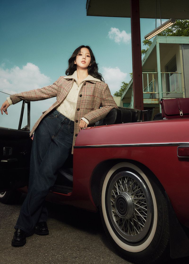 A woman in jeans and a jacket poses with a red convertible car against a sky blue backdrop with a building and some trees.