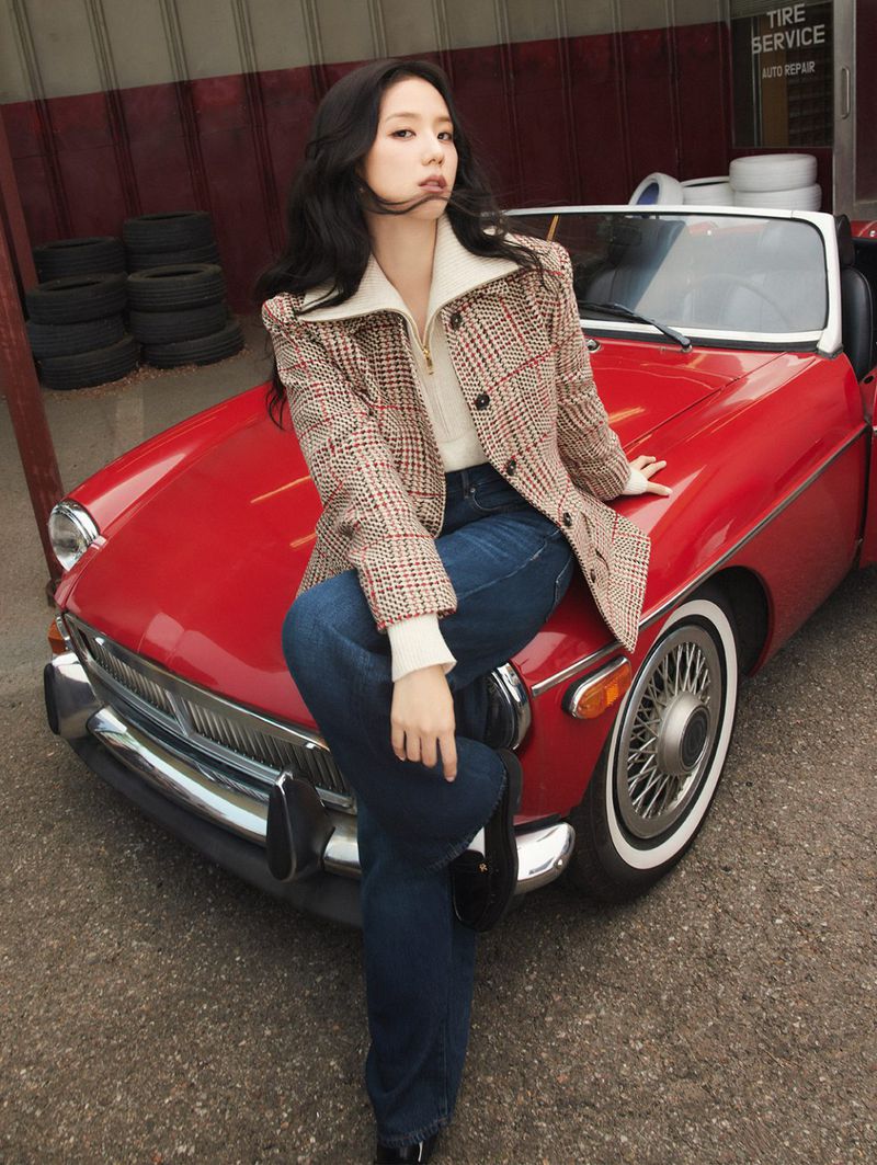 A woman with long dark hair wearing a patterned coat poses with a red convertible car in front of a building with tire repair service signage.