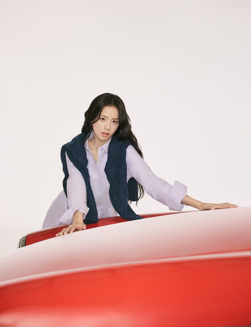 An adult woman poses in a studio setting with a red car, wearing a light purple button-down shirt and a navy blue sweater vest.